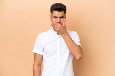 Beyazlı genç bir adam bej arka planda izole edilmiş. Ağzını eliyle kapatıyor.