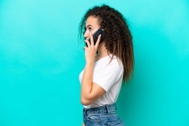 Genç bir Arap kadın mavi arka planda izole edilmiş cep telefonuyla biriyle sohbet ediyor.