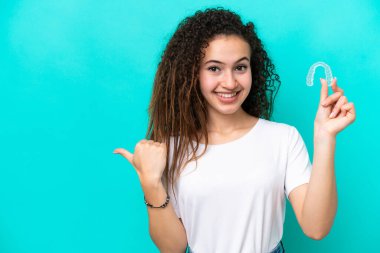 Mavi arka planda izole edilmiş görünmez diş telleri tutan genç bir Arap kadın bir ürünü sunmak için kenarı işaret ediyor.