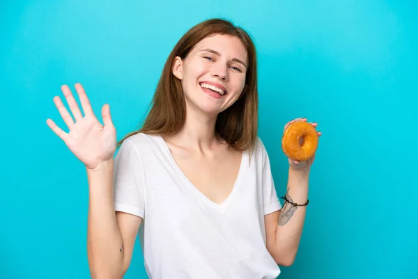 Молодая Английская Женщина Держит Пончик Изолированном Синем Фоне Приветствуя Руками — стоковое фото