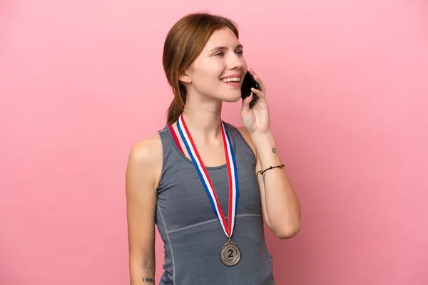 Young English woman with medals isolated on pink background keeping a conversation with the mobile phone