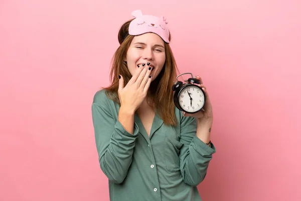 Young English woman in pajamas isolated on pink background in pajamas and holding clock with surprised expression