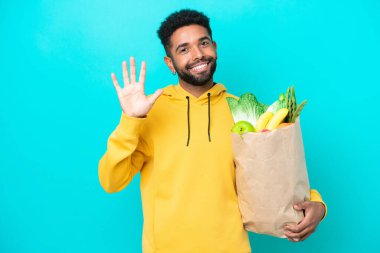 Mavi arka planda izole edilmiş bir paket yiyecek alan genç Brezilyalı adam parmaklarıyla beşi sayıyor.