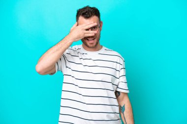 Genç Brezilyalı bir adam mavi arka planda izole edilmiş. Gözlerini elleriyle kapatıyor ve gülümsüyor.