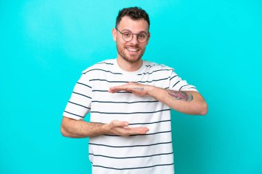 Mavi arka planda izole edilmiş genç Brezilyalı bir adam bir ilan vermek için avucunun içinde hayali bir alan tutuyor.