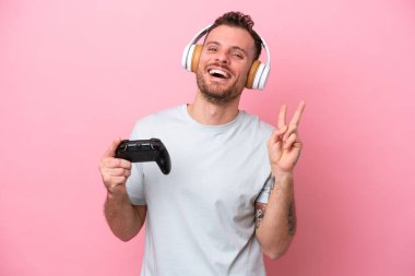 Young Brazilian man playing with video game controller isolated on pink background smiling and showing victory sign