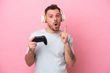 Young Brazilian man playing with video game controller isolated on pink background intending to realizes the solution while lifting a finger up