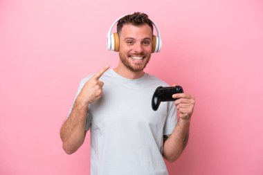 Pembe arka planda izole edilmiş bilgisayar oyunu kontrolörüyle oynayan Brezilyalı genç bir adam başparmak hareketi yapıyor.