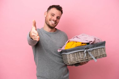 Genç Brezilyalı bir adam pembe arka planda izole edilmiş bir çamaşır sepetini tutuyor ve iyi bir anlaşma için el sıkışıyor.