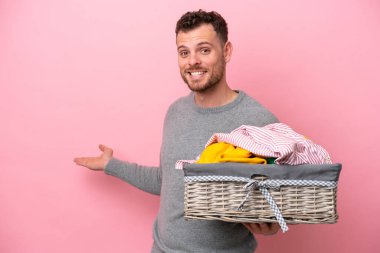 Genç Brezilyalı bir adam pembe arka planda izole edilmiş bir çamaşır sepeti tutuyor. Davet etmek için ellerini yana uzatıyor.