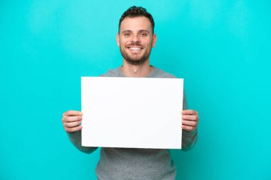 Elinde boş bir levha tutan genç Brezilyalı adam mavi arka planda izole edilmiş mutlu bir ifadeyle boş bir pankart tutuyor.