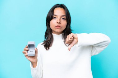 Mavi arka planda izole edilmiş bir nişan yüzüğü tutan genç İspanyol kadın olumsuz bir ifadeyle baş parmağını gösteriyor.