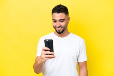 Genç Arap yakışıklı adam sarı arka planda izole edilmiş cep telefonuyla mesaj ya da e-posta gönderiyor.