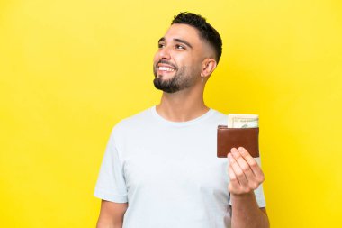 Sarı arka planda izole edilmiş cüzdan taşıyan genç bir Arap gülümserken yukarı bakıyor.