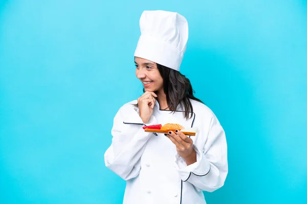 Young hispanic chef woman holding sashimi isolated on blue background looking to the side and smiling