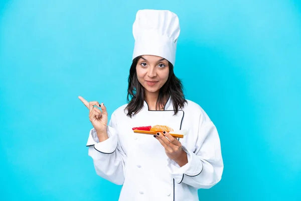 Young hispanic chef woman holding sashimi isolated on blue background pointing to the side to present a product