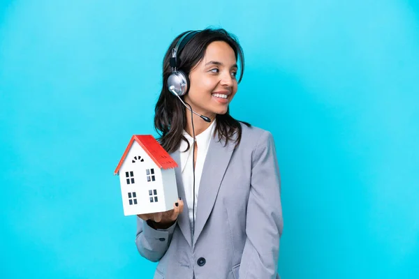 Real estate hispanic agent holding a toy house isolated on blue background looking to the side and smiling