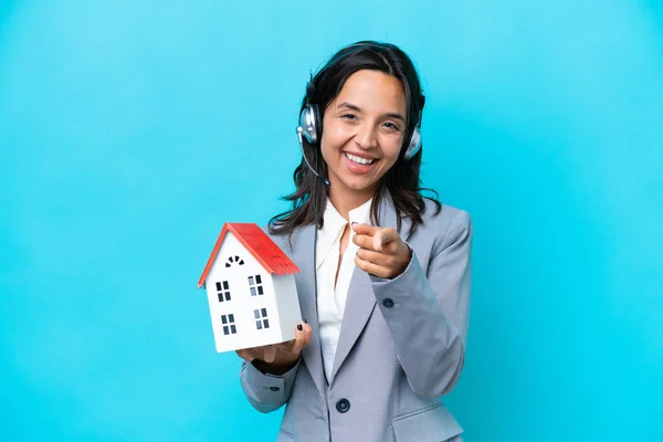 Real estate hispanic agent holding a toy house isolated on blue background pointing front with happy expression