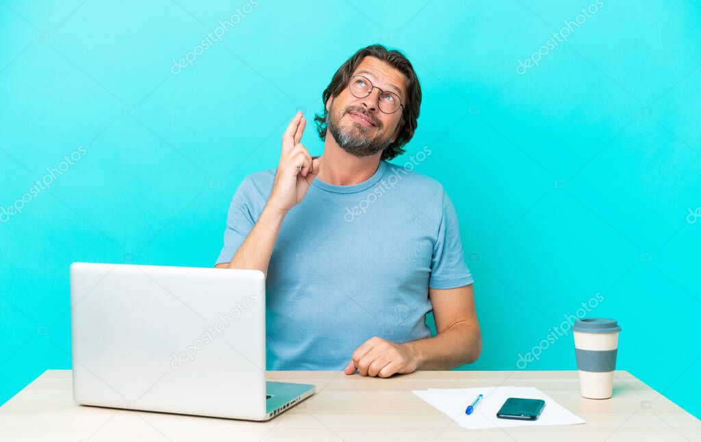Senior dutch man in a table with a laptop isolated on blue background with fingers crossing and wishing the best
