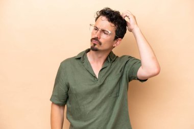 Kafkasyalı genç adam bej arka planda izole edilmiş. Kafasını kaşırken şüpheleri var.