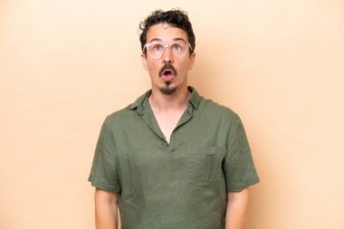 Beyazlı genç adam bej arka planda izole edilmiş yukarı bakıyor ve şaşırmış bir ifadeyle