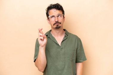 Beyaz tenli genç adam bej arka planda izole edilmiş. Parmakları çapraz ve en iyisini diliyor.