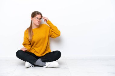 Beyaz arka planda izole bir şekilde yerde oturan beyaz bir kadın. Şüpheleri var ve kafası karışmış bir yüz ifadesi var.