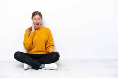Yerde oturan beyaz tenli genç kadın şaşırmış ve şok olmuş bir yüz ifadesiyle beyaz arka planda izole edilmiş.