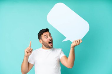 Mavi arka planda izole edilmiş yakışıklı beyaz adam boş bir konuşma balonu tutuyor ve düşünüyor.