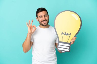 Mavi arka planda izole edilmiş yakışıklı beyaz adam elinde bir ampul simgesi tutuyor ve tamam işareti yapıyor.