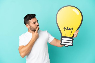 Mavi arka planda izole edilmiş yakışıklı beyaz bir adam elinde bir ampul simgesi ve şüpheleri var.