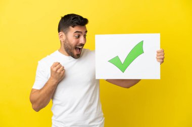 Sarı arka planda izole edilmiş yakışıklı beyaz adam elinde yeşil işaret simgesi olan bir levha tutuyor ve zaferi kutluyor.