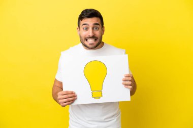 Sarı arka planda izole edilmiş yakışıklı beyaz bir adam elinde mutlu ifadeli bir ampul simgesi olan bir levha tutuyor.