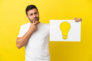 Young handsome caucasian man isolated on yellow background holding a placard with bulb icon and thinking