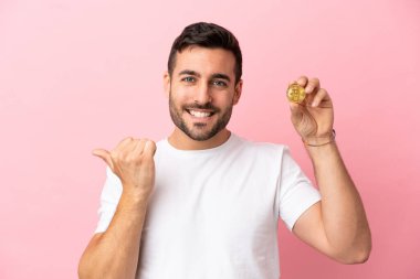 Elinde pembe arka planda izole edilmiş bir Bitcoin tutan genç adam bir ürünü sunmak için kenarı işaret ediyor.