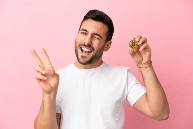 Elinde pembe arka planda izole edilmiş Bitcoin tutan genç adam gülümsüyor ve zafer işareti gösteriyor.