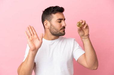 Elinde pembe arka planda izole edilmiş Bitcoin tutan genç adam dur hareketi yapıyor ve hayal kırıklığına uğruyor