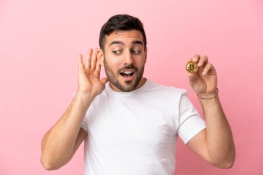 Pembe arka planda izole edilmiş bir Bitcoin tutan genç adam bir şey dinlemek için elini kulağına koydu.