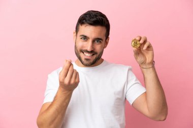 Elinde pembe arka planda izole edilmiş bir Bitcoin tutan genç adam para hareketi yapıyor.