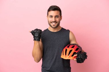 Pembe arka planda izole edilmiş genç bisikletçi bir ürünü sunmak için kenarı işaret ediyor.