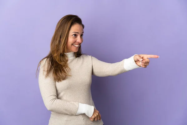 巴西中年妇女被紫色背景隔离 手指指向侧面并展示了一种产品 — 图库照片