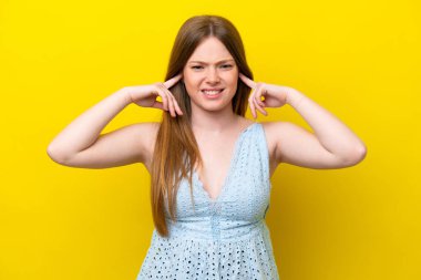 Sarı arka planda izole edilmiş genç beyaz kadın hayal kırıklığına uğramış ve kulaklarını kapatmış.