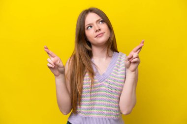 Sarı arka planda izole edilmiş genç beyaz kadın parmaklarını çapraz yapıp en iyisini diliyor.