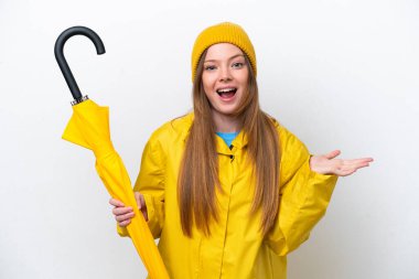 Yağmura dayanıklı ceketi ve şemsiyesi olan beyaz arka planda, yüzü şoke olmuş genç beyaz bir kadın.