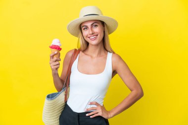 Sarışın Uruguaylı kız, elinde dondurma tutarken sarı arka planda izole edilmiş, kolları kalçasında poz veriyor ve gülümsüyor.