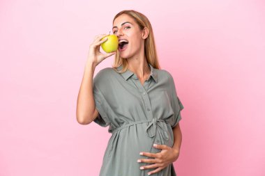 Genç Uruguaylı kadın mavi arka planda izole edilmiş hamile ve bir elma tutuyor ve yiyor.