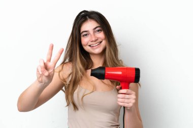 Young caucasian woman holding a hairdryer isolated on white background smiling and showing victory sign