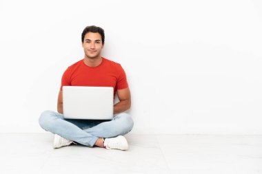 Kafkasyalı, laptoplu yakışıklı adam yerde oturmuş gülüyor.