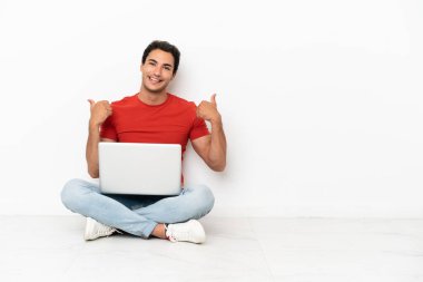 Caucasian handsome man with a laptop sitting on the floor with thumbs up gesture and smiling