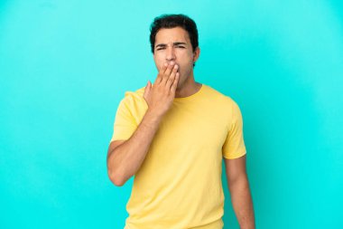 Kafkas yakışıklı adam izole edilmiş mavi arka planda esniyor ve geniş ağzını eliyle kapatıyor.